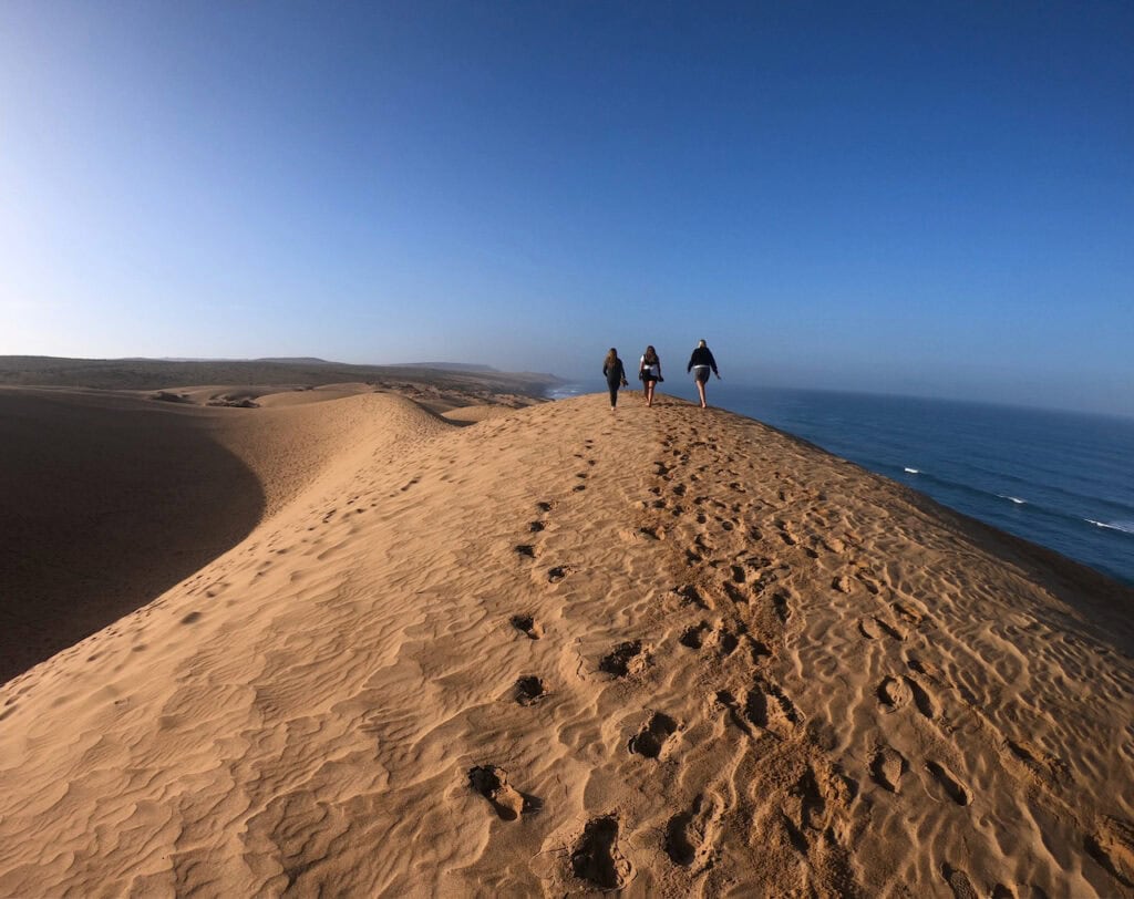 Sandklit med tre personer, der går mod horisonten ved havet, solrig dag, naturskøn udsigt.
